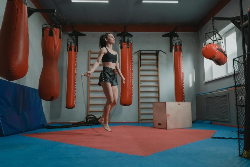 Mujer saltando a la comba en un gimnasio rodeada de varios sacos de boxeo, haciendo gala de su agilidad y resistencia.