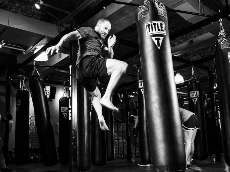 Peleador masculino saltando, preparado para patear un saco de boxeo con potencia y precisión, Luchador masculino saltando, listo para patear un saco de boxeo con potencia y precisión, mostrando cómo las artes marciales transforman la mente y el cuerpo.